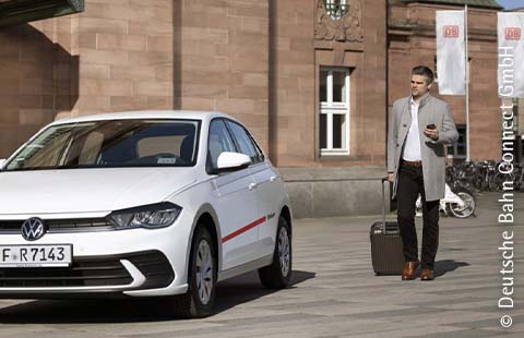 Ein Mann mit einem Rollkoffer und einem Handy in der Hand läuft vor einem Bahnhof auf ein weißes Flinkster Carsharing Fahrzeug zu.